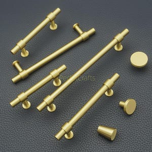 May include: A collection of gold-colored cabinet hardware, including several bar pulls and two knobs. The pulls vary in length, with a cylindrical bar and circular mounting bases. A conical knob is also present. The hardware is displayed on a dark surface.
