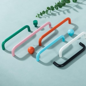 May include: A collection of colorful cabinet hardware, including green, pink, orange, blue, white, and black handles. Also shown are a green knob and two spherical knobs in orange and blue. The hardware is arranged on a light blue surface.
