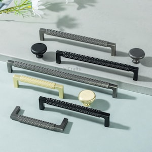 May include: A collection of cabinet hardware, including handles and knobs, in various metallic finishes. The selection features black, gold, and gray options with textured and smooth designs. The hardware is arranged on a light gray surface.