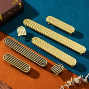 Könnte beinhalten: Eine Sammlung goldener und schwarzer Möbelbeschläge, darunter Griffe und dekorative Elemente. Die Griffe haben ein gerilltes Design und sind in verschiedenen Größen erhältlich. Die Beschläge sind auf einer blau-orangenen Oberfläche angeordnet.