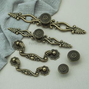 May include: Antique brass cabinet hardware set. Includes two ornate pulls, two smaller pulls, and two round knobs. The hardware features floral and scrollwork designs. The text "PerfecCrafts" is visible on the image.