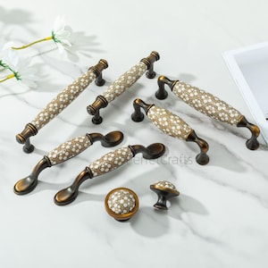 Pode incluir: Um conjunto de puxadores e puxadores decorativos para armários. Os puxadores têm um acabamento bronze com um padrão floral branco e castanho. Os puxadores têm um design correspondente. O hardware é exibido em uma superfície de mármore branco.