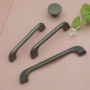 Op de afbeelding: Een set grijze kastbeslag, waaronder drie handgrepen en een ronde knop. De handgrepen hebben een gebogen ontwerp en zijn gemaakt van mat metaal. De tekst "PerfetCrafts" is zichtbaar op de onderste handgreep.