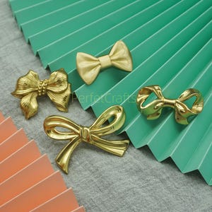 Op de afbeelding: Vier goudkleurige, strikvormige decoratieve hardware-onderdelen. De strikken variëren in ontwerp, sommige met ingewikkelde details en andere met een eenvoudiger, gestroomlijnder uiterlijk. De strikken zijn gerangschikt op een grijs oppervlak met een groen en oranje waaiervormige achtergrond.