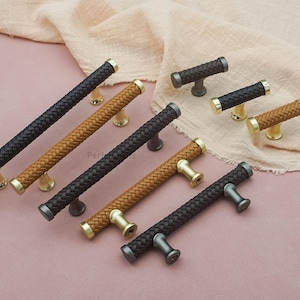 Pode incluir: Uma variedade de puxadores de armário com um design de couro trançado em preto, castanho e bege. Os puxadores têm bases e extremidades douradas, prateadas e bronze. Os puxadores estão dispostos numa superfície rosa.