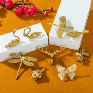 May include: A collection of gold-colored decorative figurines, including a swan, bird, dragonfly, deer antlers, butterfly, seashell, and fish. The figurines are displayed on white platforms against an orange background.