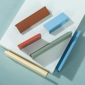 Könnte beinhalten: Eine Auswahl an farbenfrohen Schrankgriffen in verschiedenen Farbtönen, darunter Braun, Blau, Rot, Grün und Gelb. Die Griffe sind rechteckig und haben ein modernes Design. Sie sind auf einer weißen Oberfläche vor einem hellblauen Hintergrund angeordnet.