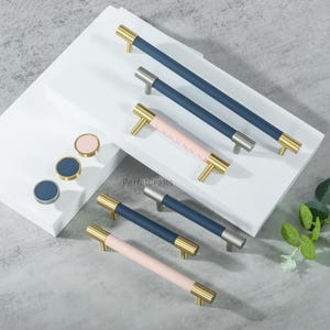 May include: A collection of cabinet hardware, including navy blue and pink leather-wrapped handles with gold and silver accents. Also, round cabinet knobs in navy blue and pink with gold and silver trim are displayed.