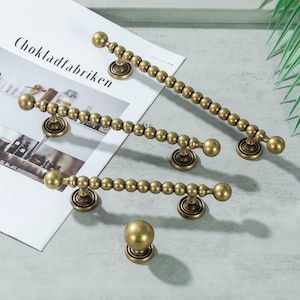 May include: A set of antique brass cabinet handles and a knob. The set includes three beaded pull handles and one round knob. The hardware is displayed on a light grey surface.