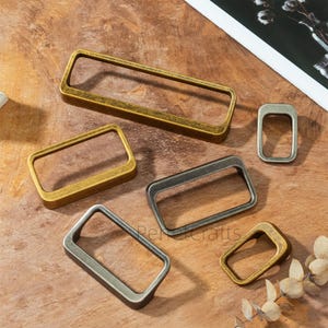 May include: An assortment of rectangular metal rings in various sizes and finishes, including gold, bronze, and silver. The rings are arranged on a wooden surface, showcasing their different colors and textures. These rings are likely used for crafting or jewelry making.