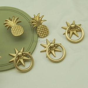 May include: Gold-colored cabinet hardware featuring pineapple and starburst designs. The pineapple knobs have textured surfaces, while the starburst knobs have a ring pull. The hardware is likely made of metal and is suitable for furniture or decor.