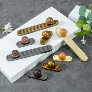 May include: A collection of decorative cabinet pulls in various metallic finishes, including gold, bronze, and gray. Each pull features a rounded knob atop a rectangular base with grooved detailing. The knobs are made of wood or metal.