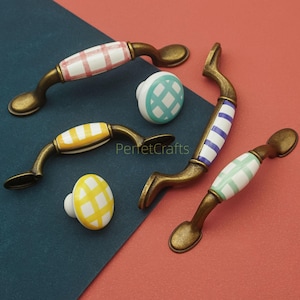 Könnte beinhalten: Eine Auswahl an dekorativen Möbelbeschlägen, darunter Griffe und Knöpfe. Die Griffe haben eine Bronzefinish mit Keramikeinsätzen in verschiedenen Mustern: rot-weißes Vichy, gelb-weißes Vichy, blau-weiße Streifen und grün-weiße Streifen.