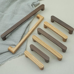 Könnte beinhalten: Eine Sammlung von Holzschrankgriffen in verschiedenen Größen und Farbtönen von Braun und Naturholz. Die Griffe haben ein rechteckiges Design mit abgerundeten Enden und sind auf einem hellgrauen Stoff angeordnet.