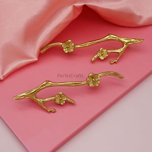 Puede incluir: Dos tiradores dorados en forma de rama con detalles de flores. Los tiradores son de metal y tienen una superficie lisa y reflectante. El fondo es una superficie rosa con tela de satén rosa.