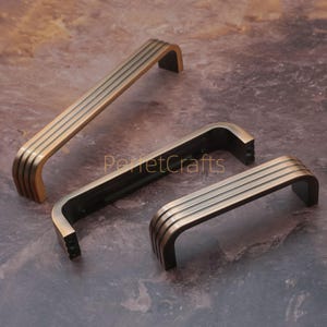 May include: Three bronze-colored cabinet pulls with a curved, rectangular design. Each pull features four horizontal grooves. The pulls are arranged on a brown, textured surface, showcasing their antique aesthetic.