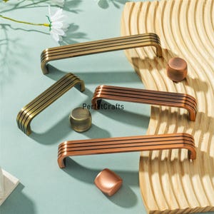 Puede incluir: Una colección de herrajes para armarios en tonos bronce y cobre, que incluyen tiradores y pomos. Los tiradores tienen un diseño curvo con tres líneas paralelas. La palabra "PerfctCrafts" está impresa en la imagen.