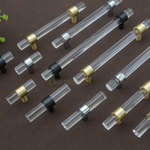 Może przedstawiać: Przezroczyste akrylowe uchwyty do szafek ze złotymi, srebrnymi i czarnymi metalowymi akcentami. Uchwyty są cylindryczne i ułożone na brązowej powierzchni. Okucia przeznaczone są do mebli i dekoracji wnętrz.