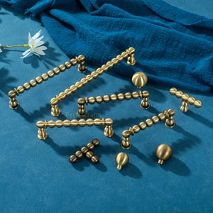 May include: Various brass cabinet handles and knobs displayed on a blue surface. The handles have a beaded design, and the knobs are spherical. The hardware is in different sizes and shapes, suitable for furniture.