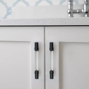 May include: Close-up of white cabinet doors with two clear and black cabinet pulls. The pulls are cylindrical with black caps and bases. The cabinet doors are plain white, and the countertop is a speckled gray.
