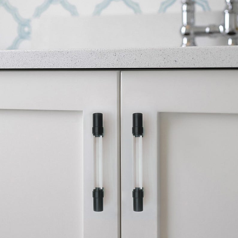 May include: Close-up of white cabinet doors with two clear and black cabinet pulls. The pulls are cylindrical with black caps and bases. The cabinet doors are plain white, and the countertop is a speckled gray.