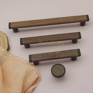 May include: A collection of wooden cabinet hardware, including three rectangular pulls and one round knob. The pulls and knob feature a dark wood grain center with black end caps and bases. The text "PerfectCrafts" is visible on the middle pull.