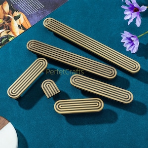 May include: A collection of brass-toned cabinet pulls and knobs with a grooved design. The hardware pieces vary in size and shape, including long rectangular pulls and smaller, rounded knobs. The text "PerfektCrafts" is visible on the surface.