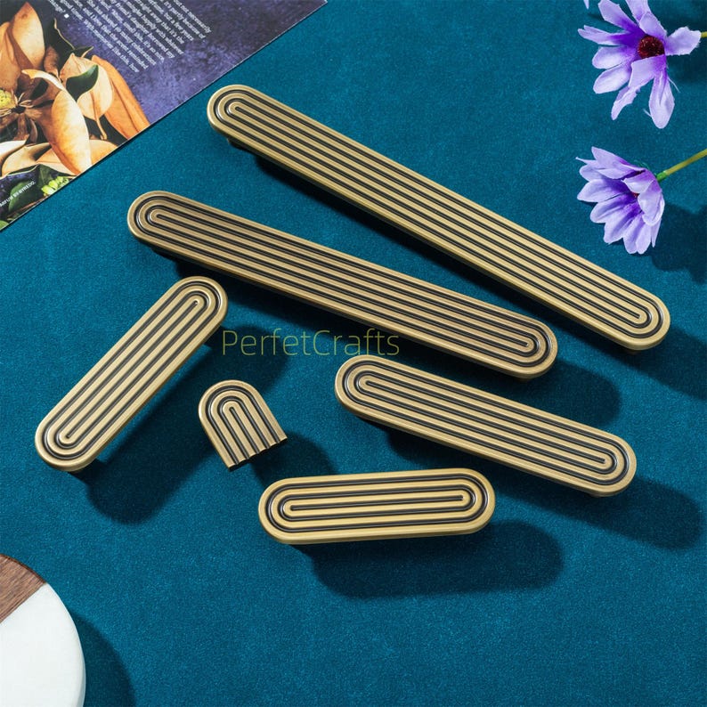 May include: A collection of brass-toned cabinet pulls and knobs with a grooved design. The hardware pieces vary in size and shape, including long rectangular pulls and smaller, rounded knobs. The text "PerfektCrafts" is visible on the surface.