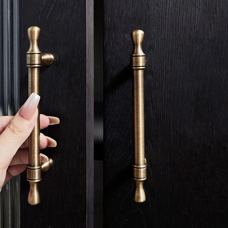 May include: Antique brass cabinet pulls with a vertical design. The pulls have a cylindrical body with decorative detailing and a rounded top and bottom. One pull is being held by a hand, showing the mounting hardware.