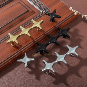 May include: A collection of star-shaped cabinet hardware in gold, black, and silver finishes. The hardware is displayed on a wooden cabinet door and a brown surface. The hardware includes both single knobs and double-knob pulls.