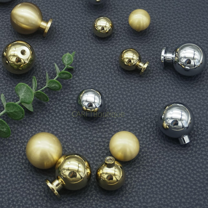 Puede incluir: Una variedad de pomos y tiradores dorados y plateados para armarios. Los pomos son esf&eacute;ricos con acabado dorado o plateado. Algunos tienen una peque&ntilde;a base redonda. Los pomos est&aacute;n dispuestos sobre una superficie gris oscuro con una ramita de hojas verdes.