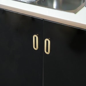 May include: Close-up of a black cabinet with two gold-coloured, oval-shaped handles. The handles have a grooved design and are mounted on the cabinet doors. The cabinet is part of a kitchen or bathroom setup.