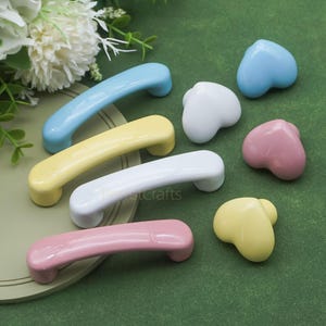 May include: A collection of colorful cabinet hardware, including pastel-colored handles and heart-shaped knobs. The handles are in blue, yellow, white, and pink. The knobs are white, light blue, pink, and yellow. The hardware is arranged on a green surface.