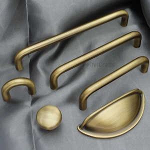 K&ouml;nnte beinhalten: Eine Sammlung von antiken Messing-Schrankbeschl&auml;gen, darunter mehrere Griffleisten, ein Knopf und ein Muschelgriff. Die Beschl&auml;ge haben eine warme, gealterte Oberfl&auml;che und sind auf einem grauen Stoff angeordnet. F&uuml;r Schr&auml;nke und Schubladen.