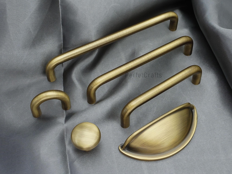 K&ouml;nnte beinhalten: Eine Sammlung von antiken Messing-Schrankbeschl&auml;gen, darunter mehrere Griffleisten, ein Knopf und ein Muschelgriff. Die Beschl&auml;ge haben eine warme, gealterte Oberfl&auml;che und sind auf einem grauen Stoff angeordnet. F&uuml;r Schr&auml;nke und Schubladen.