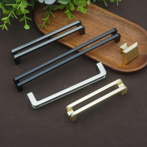 Pode incluir: Uma coleção de ferragens para armários em vários acabamentos metálicos. A seleção inclui puxadores de gaveta em preto fosco, prata e dourado e um puxador de armário dourado. O hardware é exibido em uma bandeja de madeira sobre um fundo marrom.