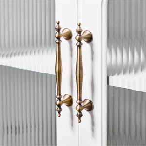 Może przedstawiać: Dwa antyczne mosiężne uchwyty do szafek z ozdobnymi detalami. Uchwyty są zorientowane pionowo, z długim, smukłym korpusem i dekoracyjnymi zwieńczeniami. Są zamontowane na białych drzwiach szafki, z teksturowanym szklanym panelem w tle.