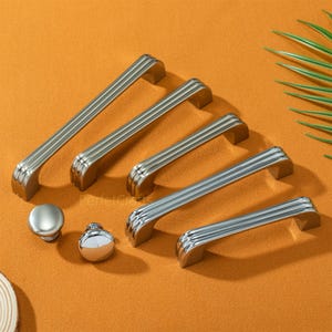 Puede incluir: Una colección de herrajes plateados para armarios, que incluye varios tiradores largos y acanalados y dos estilos de pomo. Los herrajes están dispuestos sobre una superficie naranja, mostrando la variedad de diseños disponibles.