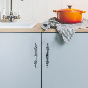 Może przedstawiać: Pomarańczowo-czerwony emaliowany żeliwny garnek holenderski z ciemnobrązową pokrywką spoczywa na pasiastym ręczniku kuchennym. Garnek holenderski stoi na drewnianym blacie nad jasnoniebieskimi drzwiami szafki z ozdobnymi srebrnymi uchwytami. Biały zlew i kran są w tle.