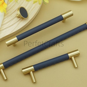 Puede incluir: Un conjunto de herrajes para armarios en azul marino y dorado, que incluye tres tiradores y un pomo. Los tiradores tienen un centro azul marino texturizado y extremos dorados. El pomo tiene un centro azul marino y una base dorada. Los herrajes se muestran sobre una superficie clara.