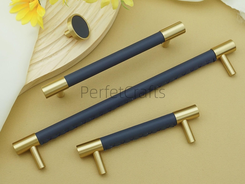 Puede incluir: Un conjunto de herrajes para armarios en azul marino y dorado, que incluye tres tiradores y un pomo. Los tiradores tienen un centro azul marino texturizado y extremos dorados. El pomo tiene un centro azul marino y una base dorada. Los herrajes se muestran sobre una superficie clara.