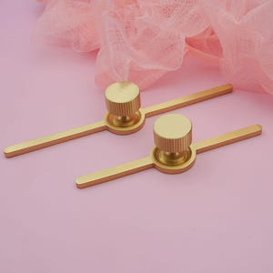 Puede incluir: Dos tiradores dorados para armarios con un dise&ntilde;o moderno. Cada tirador presenta un pomo cil&iacute;ndrico con ranuras verticales, conectado a una barra plana. Los tiradores est&aacute;n colocados sobre un fondo rosa con una tela suave y drapeada.