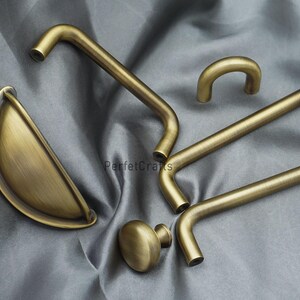 K&ouml;nnte beinhalten: Eine Auswahl an antiken Messing-Schrankbeschl&auml;gen, darunter Griffe und ein Knauf. Die Beschl&auml;ge haben einen warmen, goldenen Farbton und ein klassisches Design. Die Teile sind auf einem grauen Stoffhintergrund angeordnet.