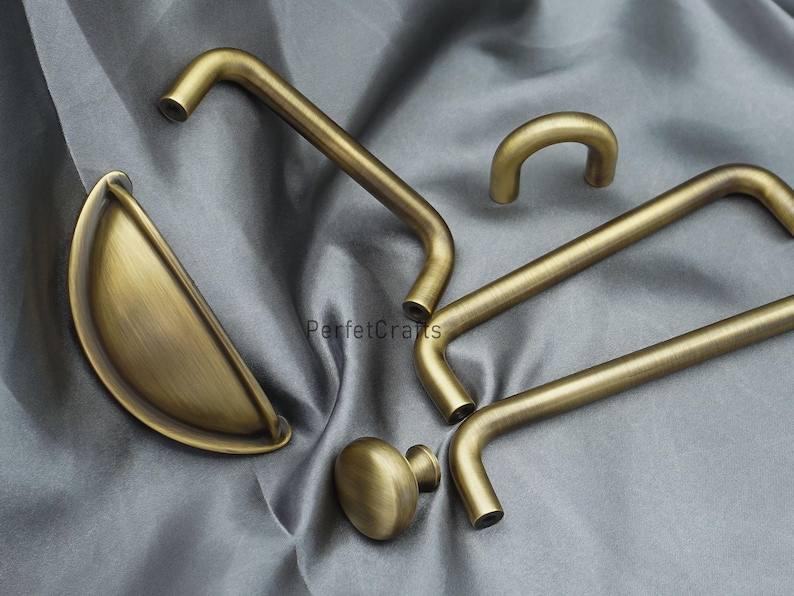 K&ouml;nnte beinhalten: Eine Auswahl an antiken Messing-Schrankbeschl&auml;gen, darunter Griffe und ein Knauf. Die Beschl&auml;ge haben einen warmen, goldenen Farbton und ein klassisches Design. Die Teile sind auf einem grauen Stoffhintergrund angeordnet.