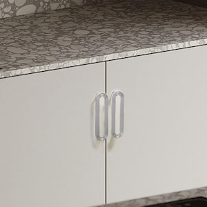 May include: A white cabinet with two silver, oval-shaped handles. The handles are vertically aligned and have a double-loop design. The cabinet doors are closed, and the countertop above is a speckled grey.