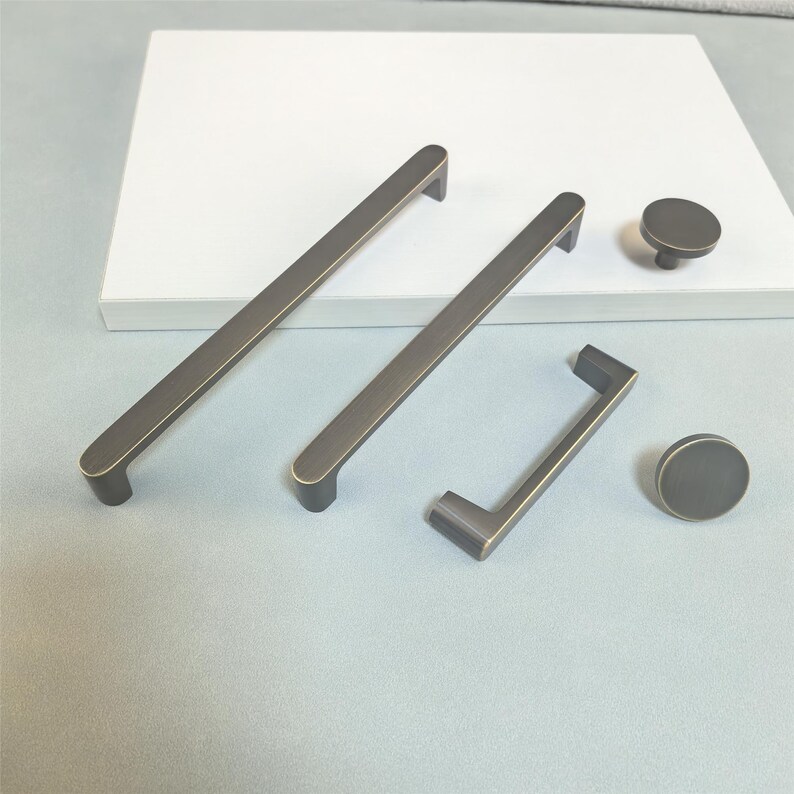 May include: A selection of bronze-coloured cabinet hardware, comprising two long bar pulls, a shorter pull, and two round knobs. The hardware is displayed on a light grey surface with a white rectangular object in the background. The hardware has a modern, minimalist design.