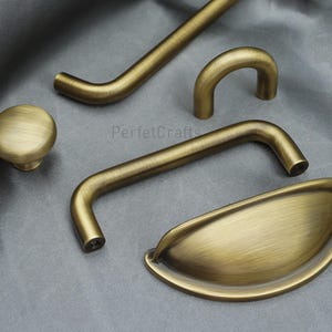 K&ouml;nnte beinhalten: Eine Auswahl an antiken Messing-Schrankbeschl&auml;gen, darunter ein Knopf, ein Muschelgriff und verschiedene Griffstile. Die Beschl&auml;ge haben eine warme, gealterte Oberfl&auml;che, ideal f&uuml;r Restaurierungsprojekte.