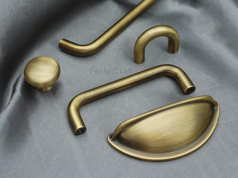 K&ouml;nnte beinhalten: Eine Auswahl an antiken Messing-Schrankbeschl&auml;gen, darunter ein Knopf, ein Muschelgriff und verschiedene Griffstile. Die Beschl&auml;ge haben eine warme, gealterte Oberfl&auml;che, ideal f&uuml;r Restaurierungsprojekte.