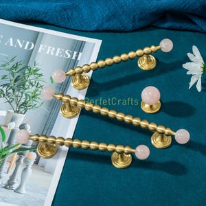 Puede incluir: Tiradores de armario de latón con detalles de piedras preciosas rosas. Los tiradores tienen un diseño de cuentas y están montados sobre bases redondas. También se ve un pomo de armario a juego. Fondo verde azulado oscuro.