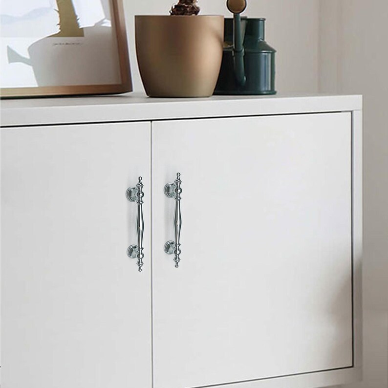 Może przedstawiać: Dwa srebrne uchwyty do szafek z ozdobnymi detalami zamontowane na białych drzwiach szafki. Uchwyty mają pionową konstrukcję z elementami dekoracyjnymi u g&oacute;ry i na dole, dodając elegancji meblom.